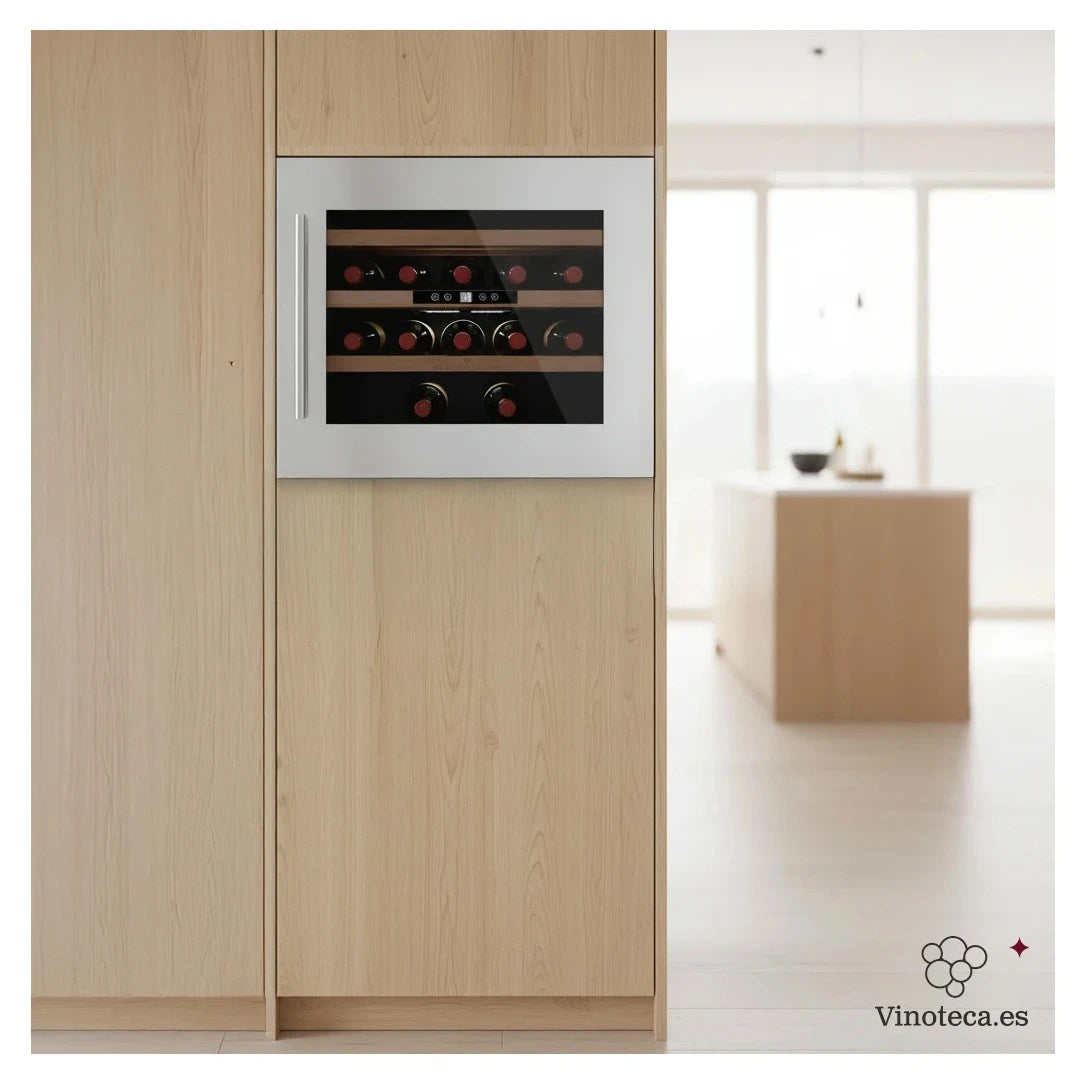 Cave de vinho Les Petits Champs CAVCI24 encastrada com moldura em aço inoxidável, integrada num móvel de madeira clara, com capacidade para 24 garrafas distribuídas em prateleiras de madeira, numa cozinha moderna e luminosa.